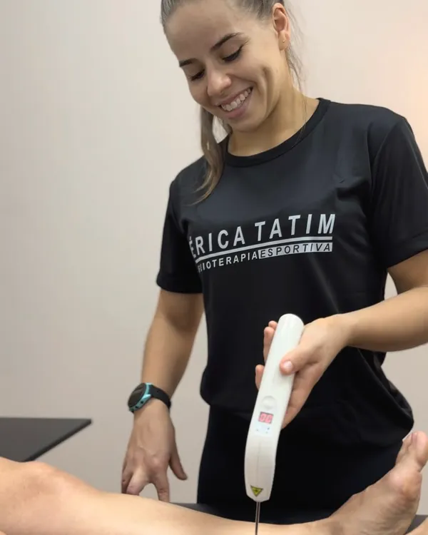 Fisioterapeuta sorrindo enquanto aplica laser terapêutico na perna de uma paciente deitada na maca