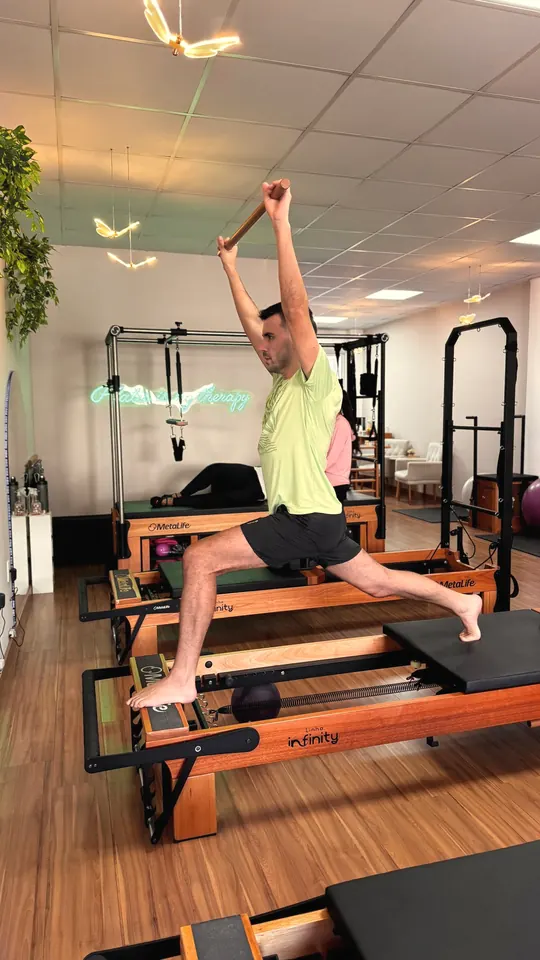 Aluno em avanço sobre o reformer, com os braços estendidos segurando um bastão